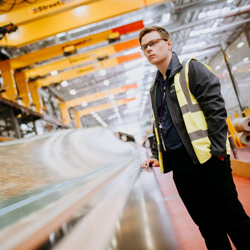 A young man in a high vis vest and safety goggles looks across an assembly line in a brightly lit factory