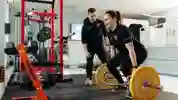 Two students in a gym, one is lifting a barbell whilst the other is spotting her