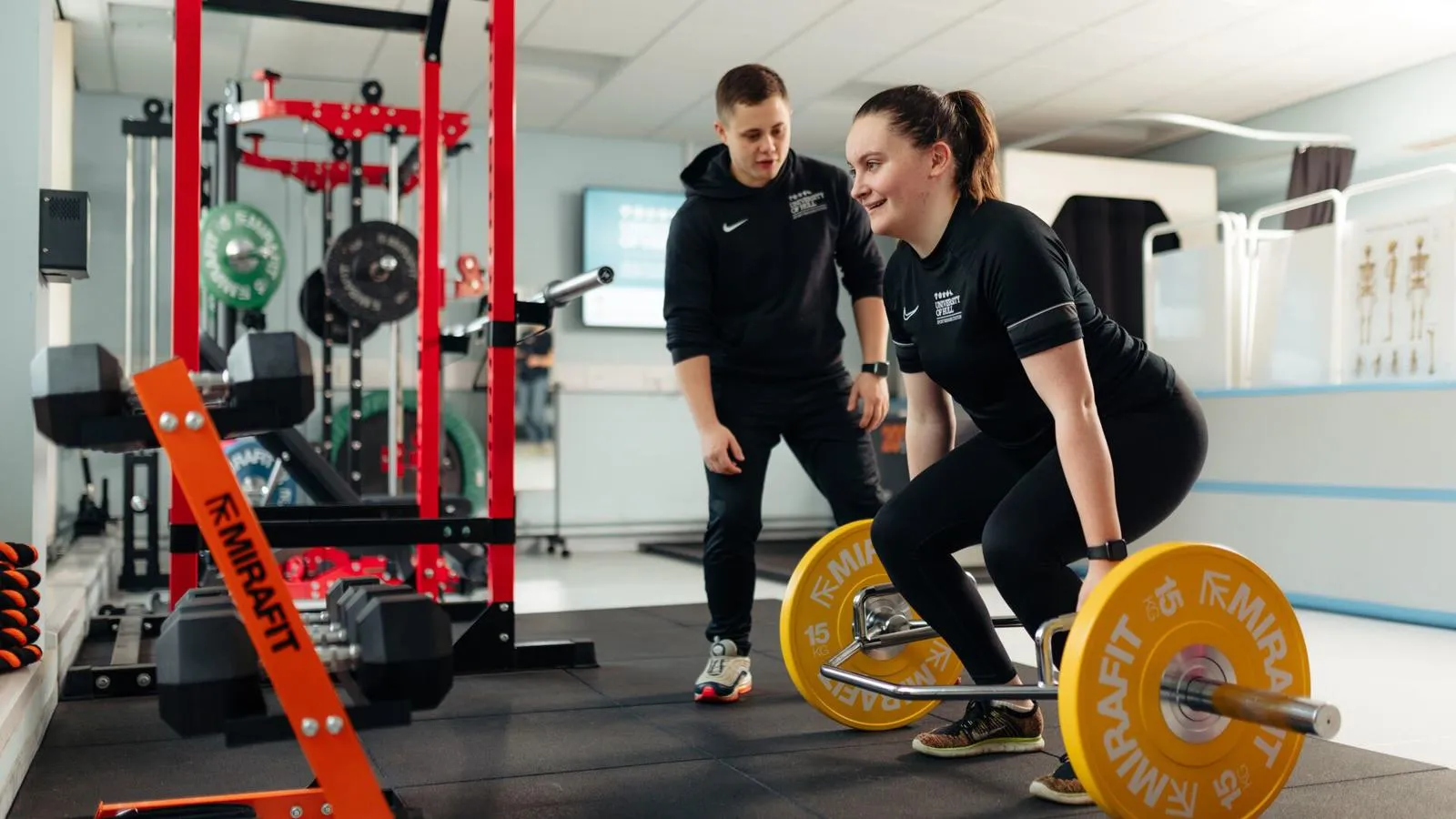 Two students in a gym, one is lifting a barbell whilst the other is spotting her