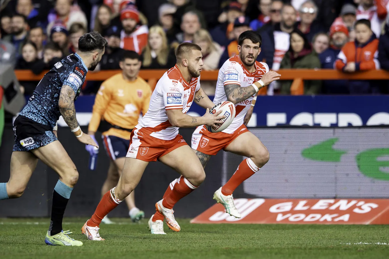 Two Hull KR rugby players run with the ball on a field during a match, while an opponent in a blue uniform approaches.