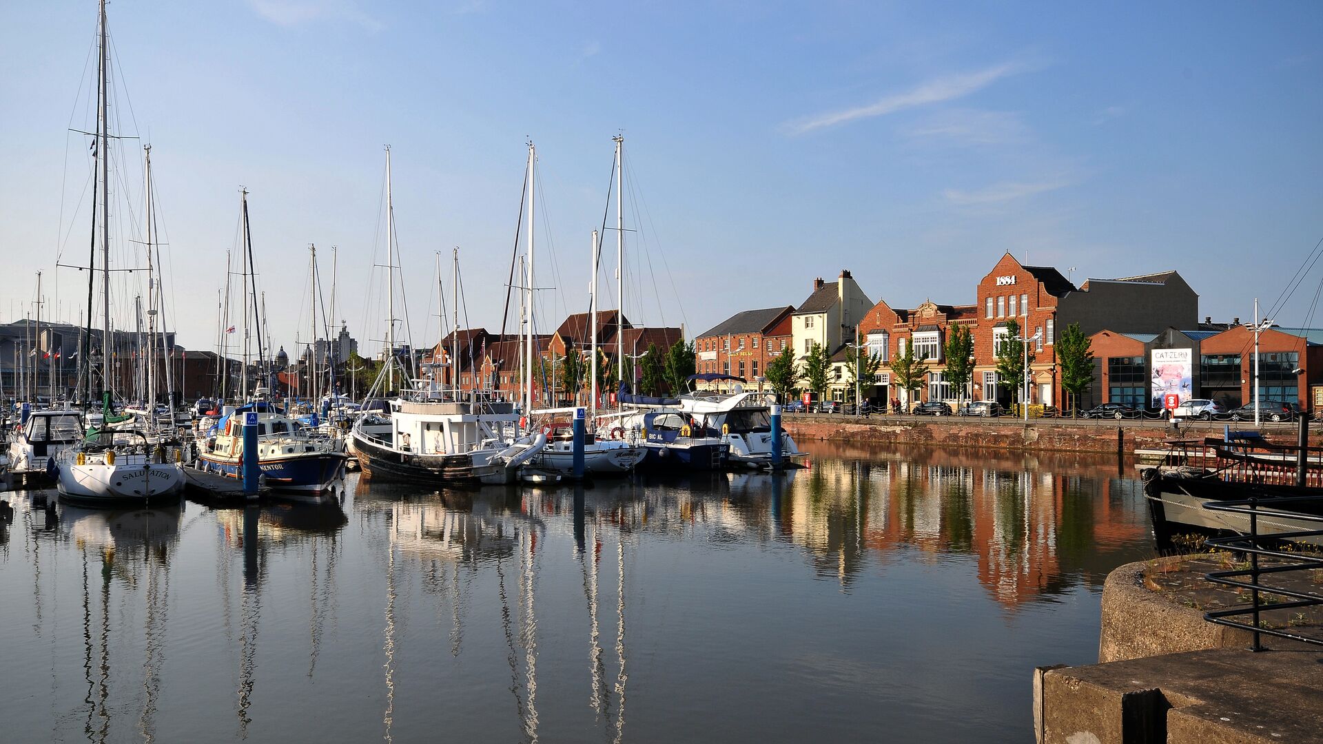 Boats docked in a marina with calm water and buildings lining the waterfront under a clear sky.