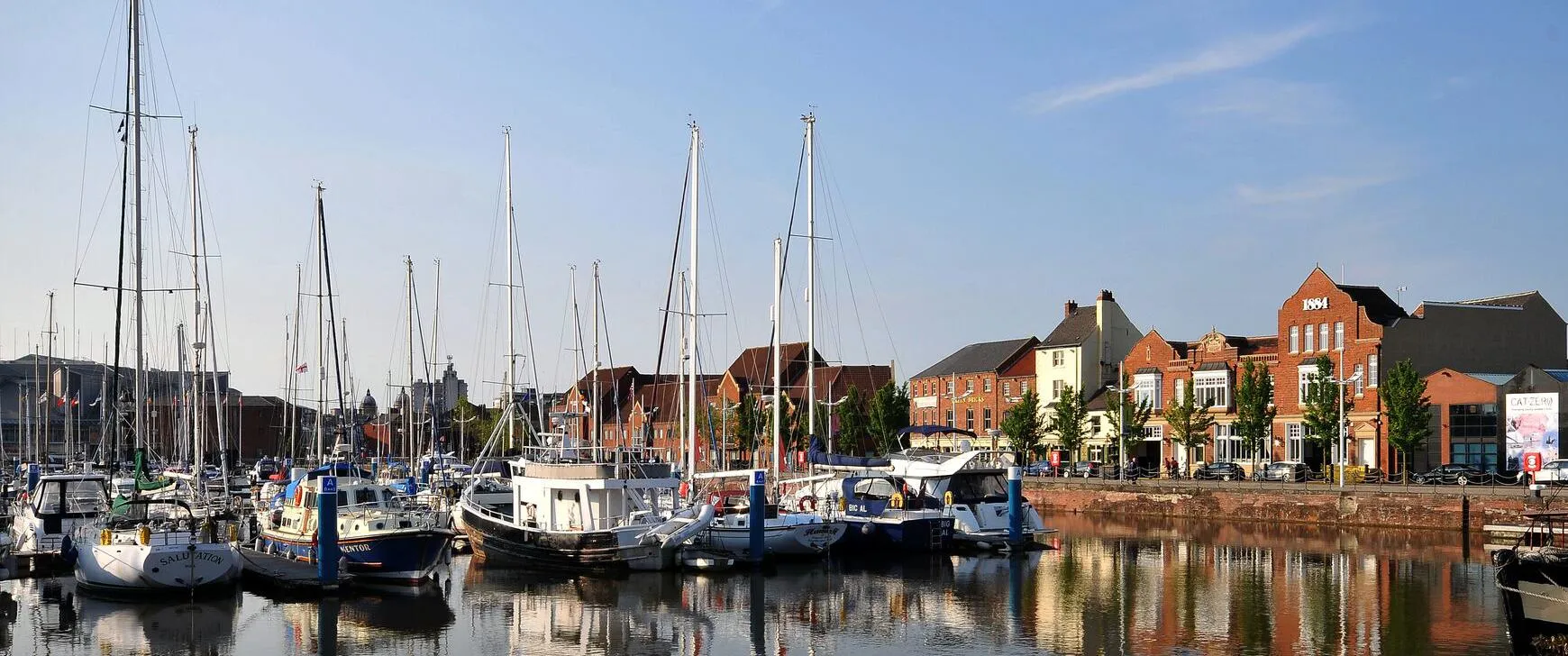Boats docked in a marina with calm water and buildings lining the waterfront under a clear sky.