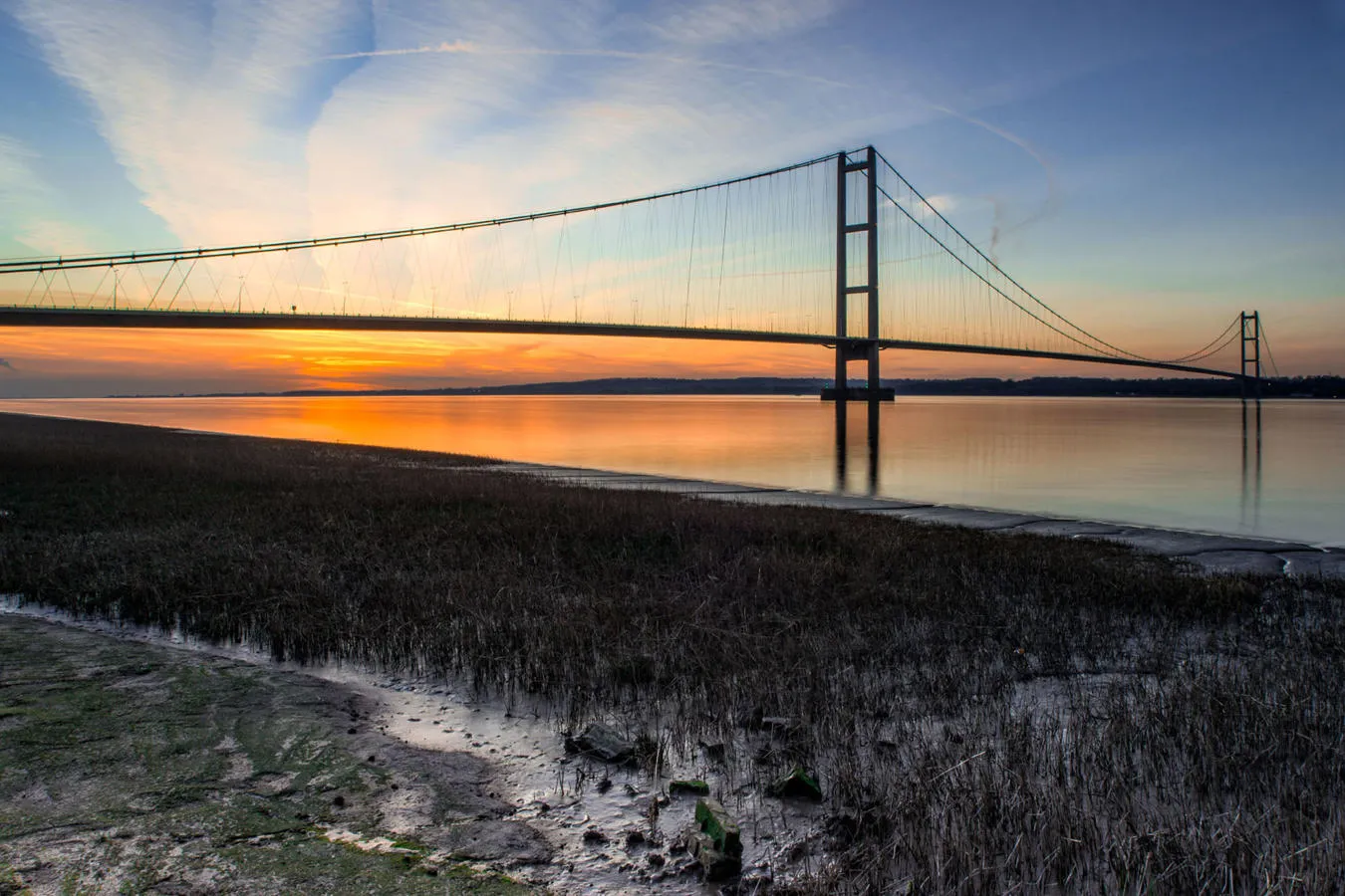 Humber Bridge at sunset and the foreground showing muddy, grassy riverbank.