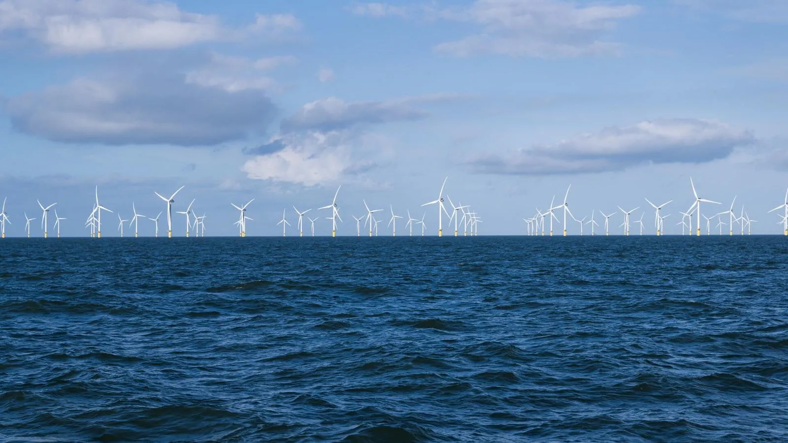 Numerous offshore wind turbines on the horizon under a cloudy blue sky with a choppy sea in the foreground.