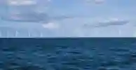 Numerous offshore wind turbines on the horizon under a cloudy blue sky with a choppy sea in the foreground.