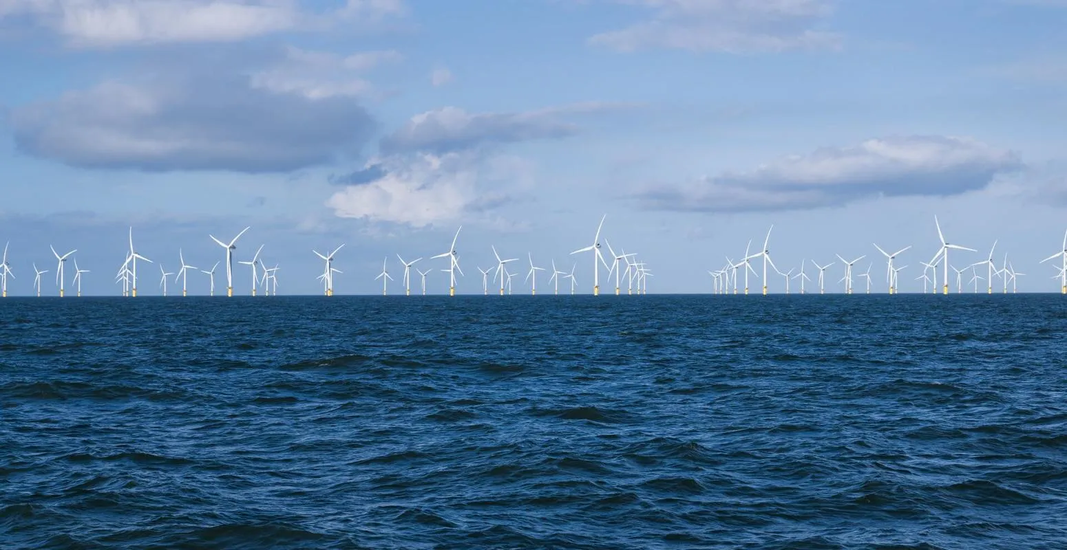 Numerous offshore wind turbines on the horizon under a cloudy blue sky with a choppy sea in the foreground.