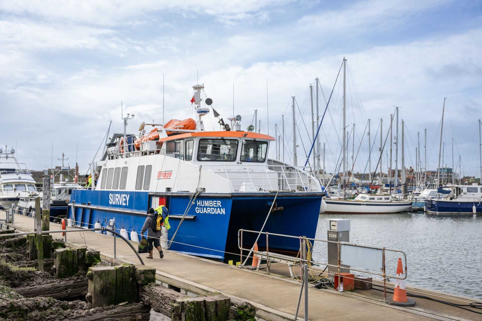 A blue and white passenger boat docked at a marina, with sailboats and masts visible in the background.
