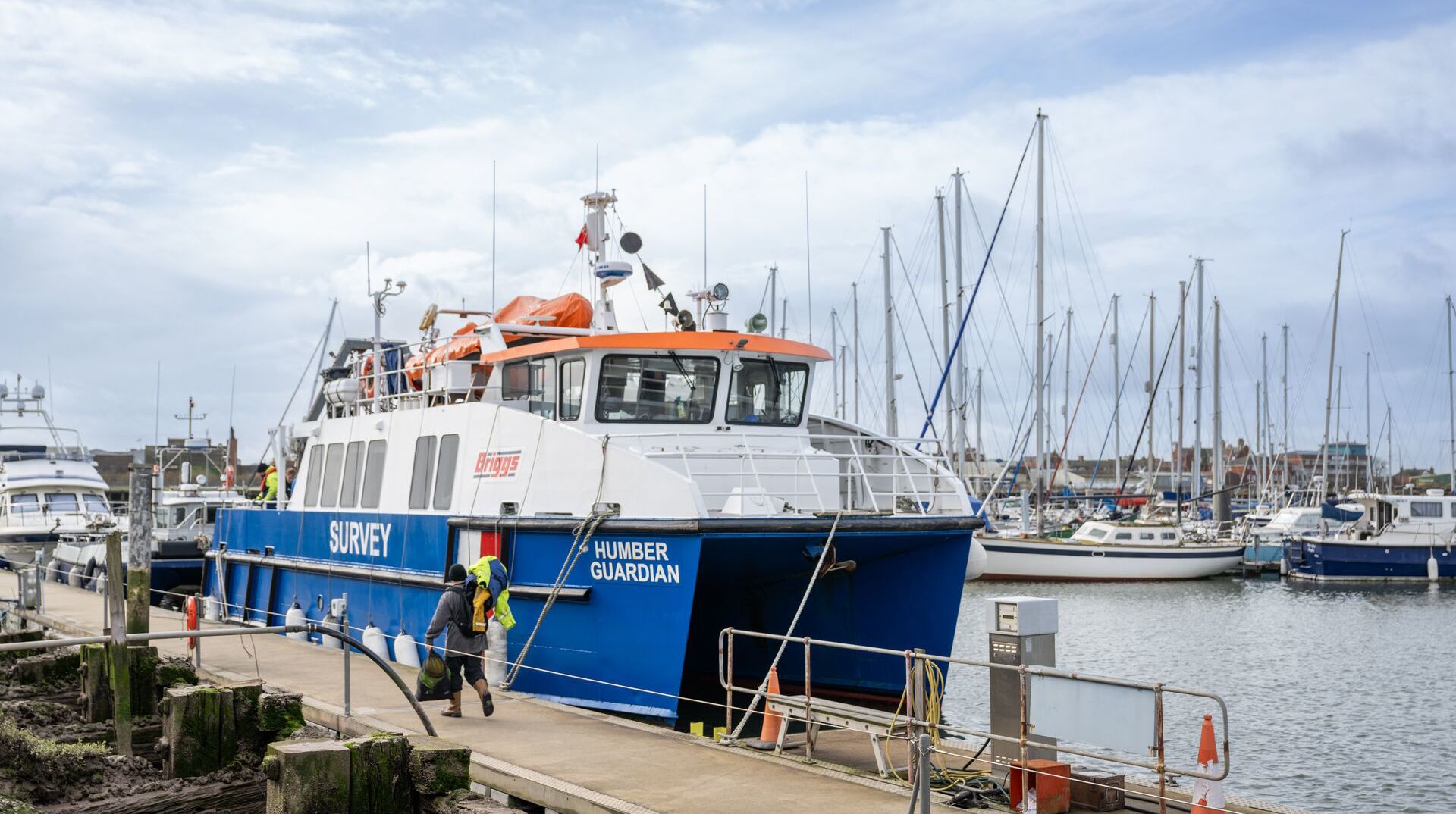 A blue and white passenger boat docked at a marina, with sailboats and masts visible in the background.