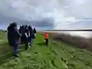A group of people walk on a grassy river bank, led by a person in a bright orange vest. 