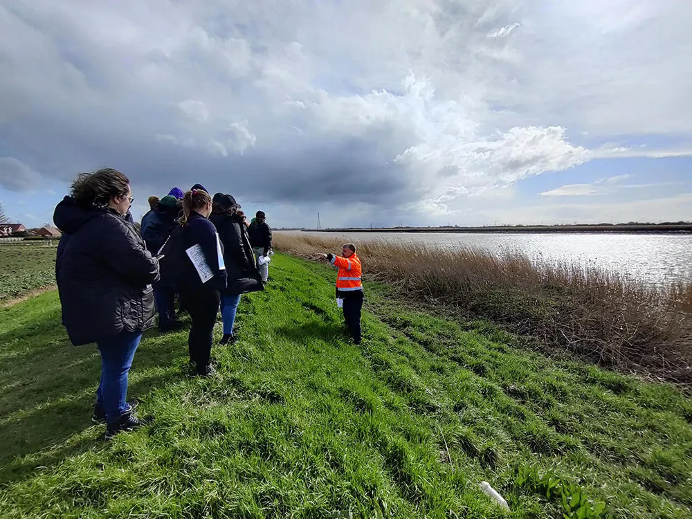 A group of people walk on a grassy river bank, led by a person in a bright orange vest. 