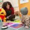 A teacher smiles at a baby while playing with a parrot puppet in a classroom