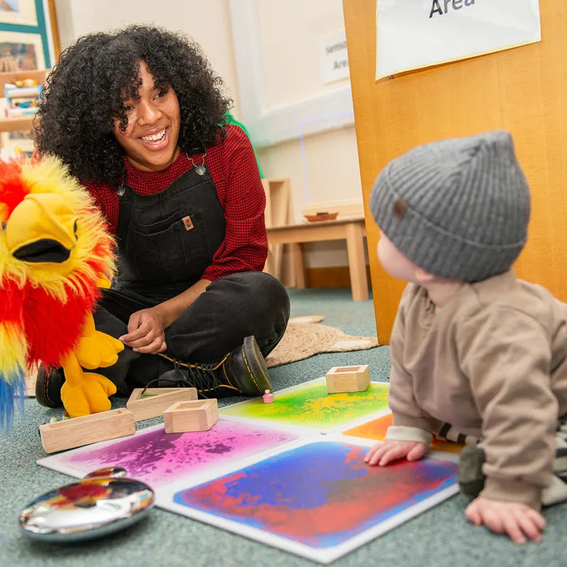 A teacher smiles at a baby while playing with a parrot puppet in a classroom
