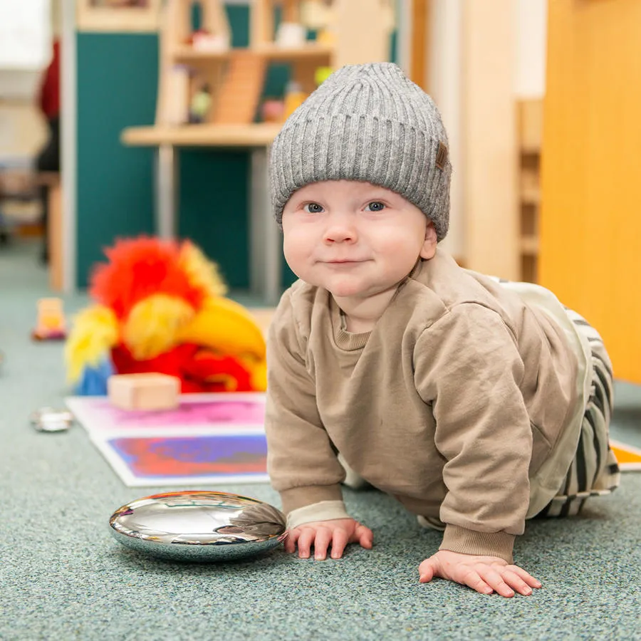 A baby in a grey hat crawls on the carpet smiling at the camera with colourful toys behind