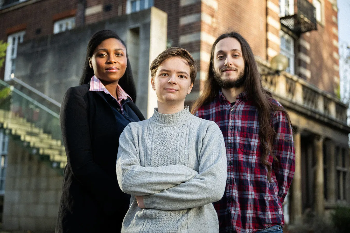 Three young adults stand together in front of a large red-brick building