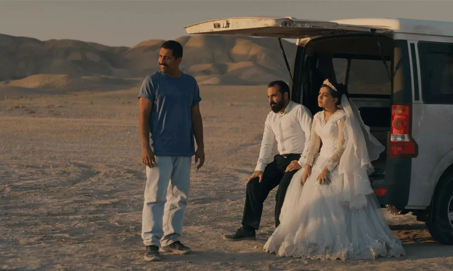 A bride and groom sit in the back of an open boot in a desert landscape