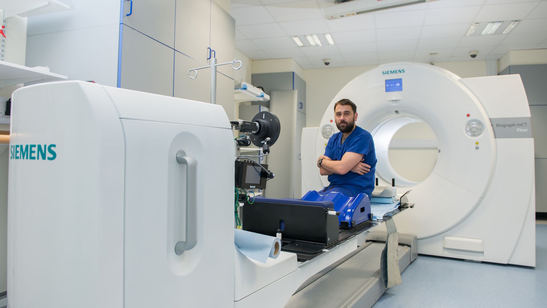 A medical professional in blue scrubs casually sits on a high tech scanning machine