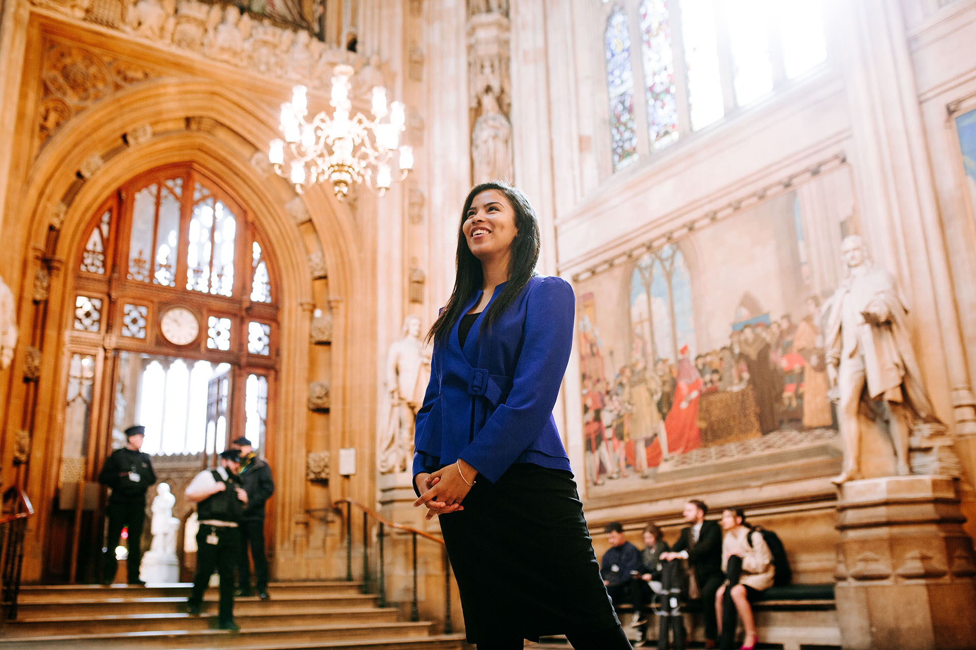 A young woman stands confidently in a historic building looking outwards with crowds in the background.