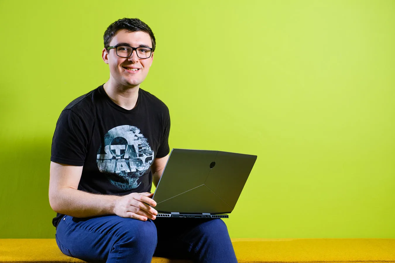 A student seated on a bench in front of a green background holds an open laptop
