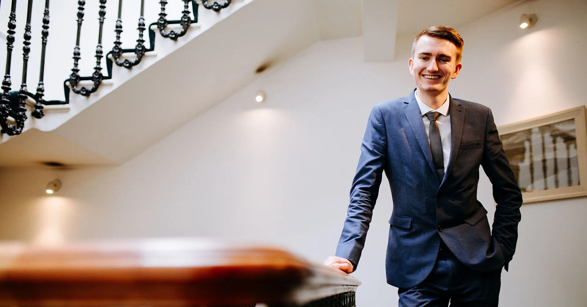 A happy young man in a smart suit stands at the top of an ornate staircase with his hand in his pocket