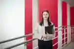 A young woman poses in front of a red wall, holding a folder, with a focused expression on her face.