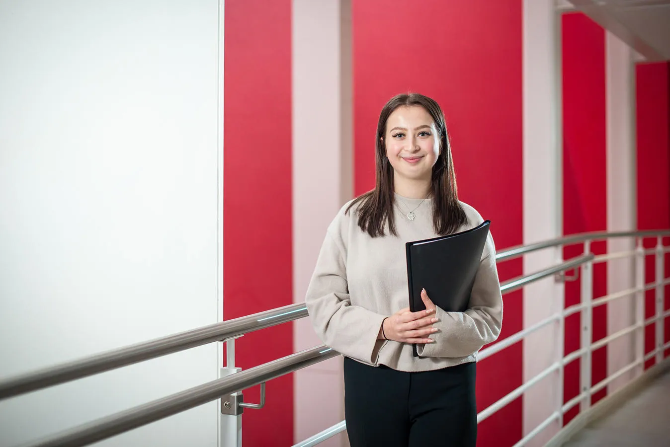 A young woman poses in front of a red wall, holding a folder, with a focused expression on her face.