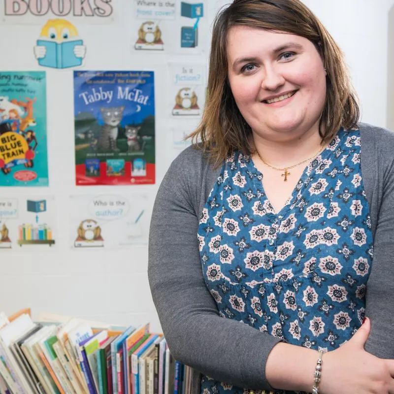 A student teacher smiles at the camera in front of a book display