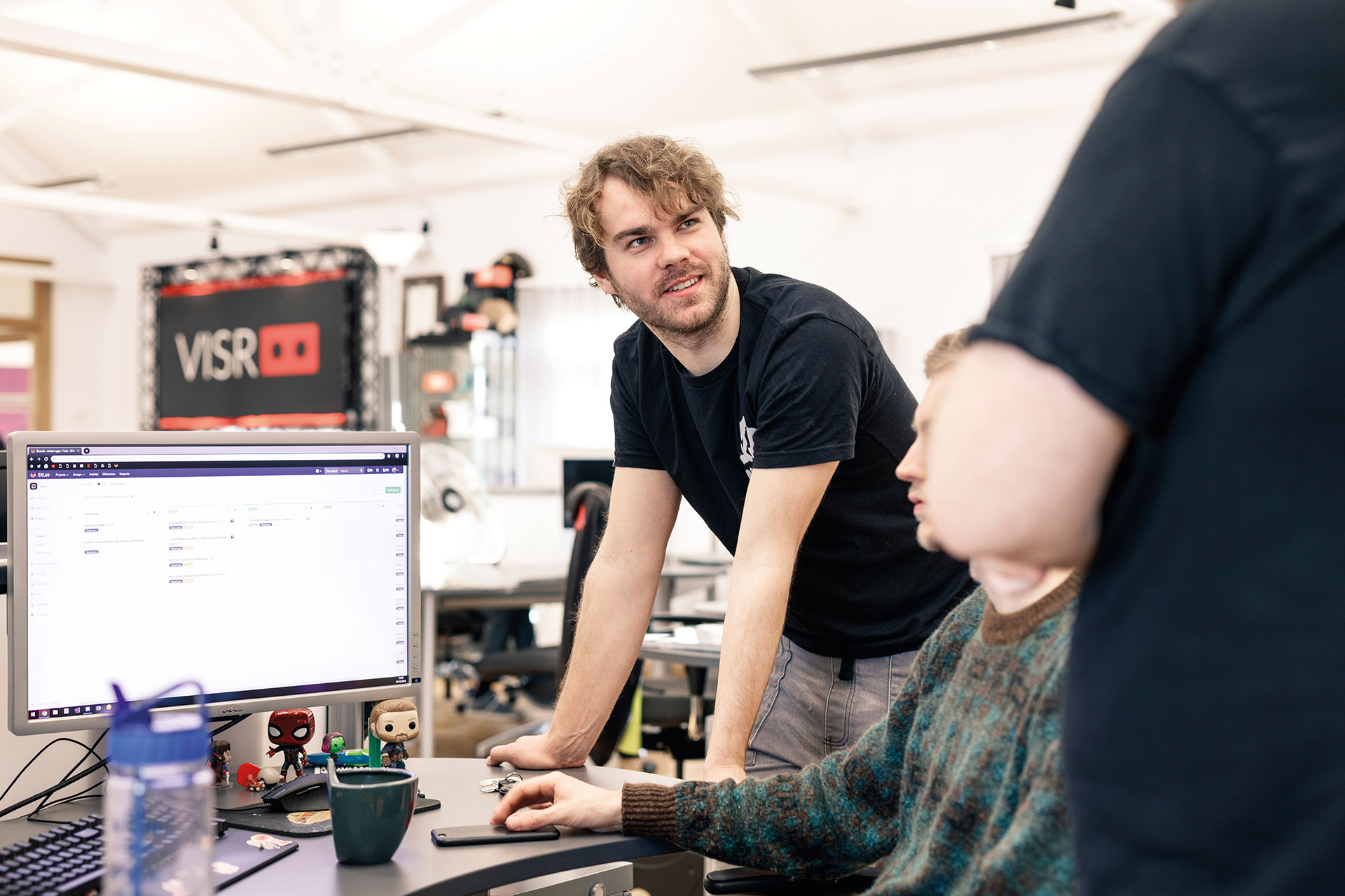 A man leans on a computer desk and engages in conversation in busy office with white walls