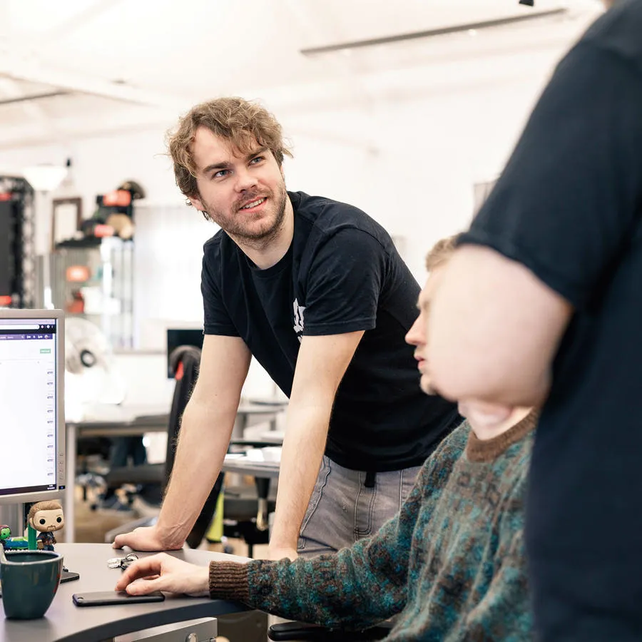A man leans on a computer desk and engages in conversation in busy office with white walls