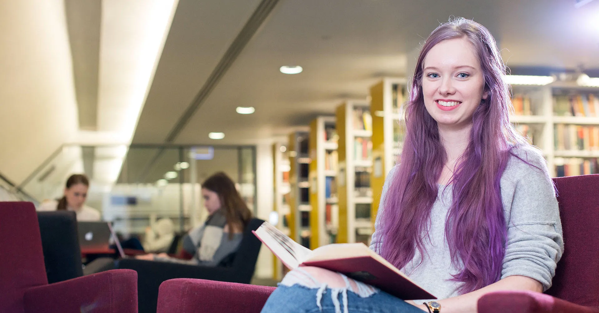 Smiling student sitting in a library with a book open on her lap