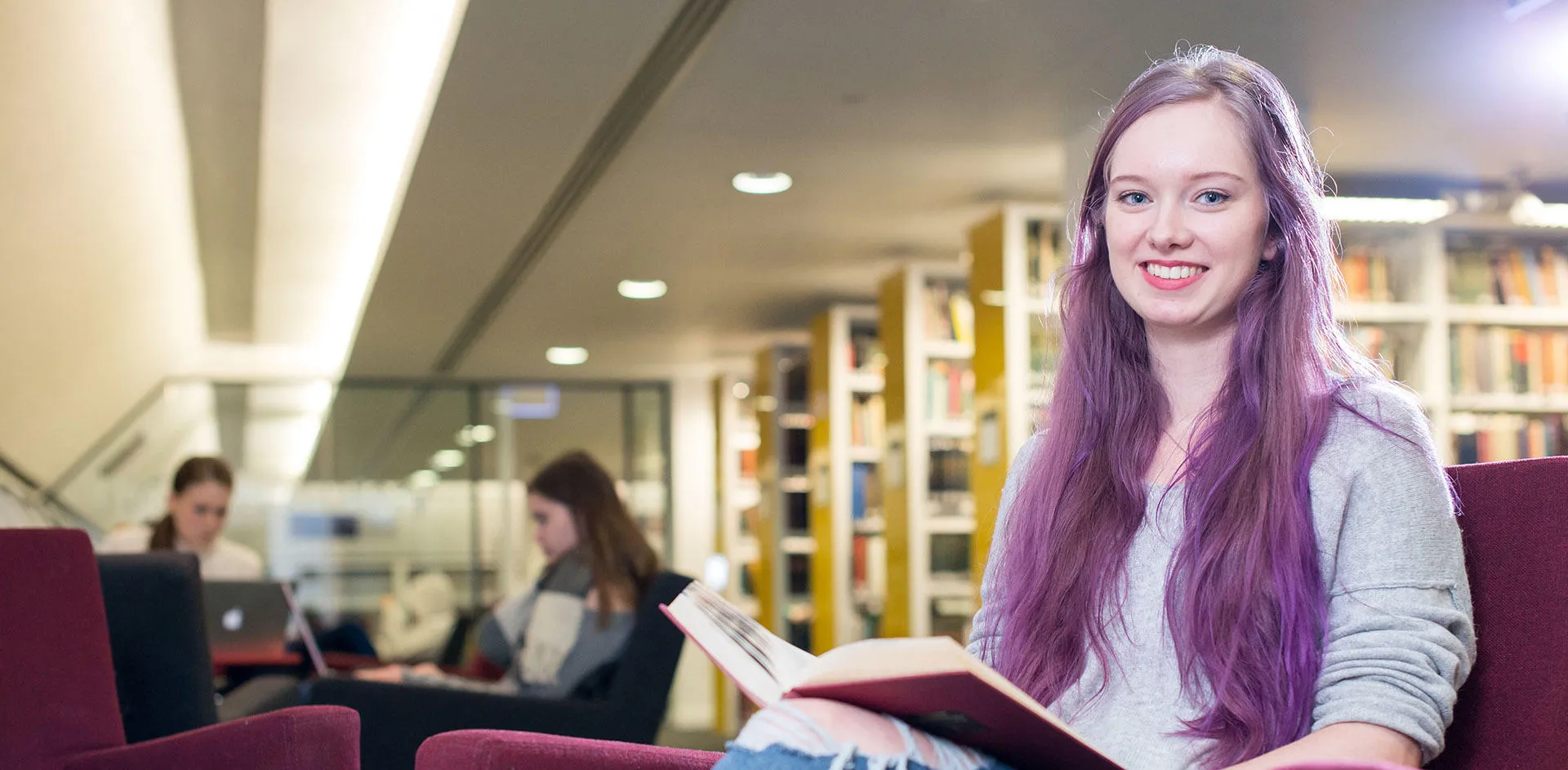 Smiling student sitting in a library with a book open on her lap