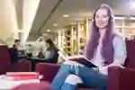 Smiling student sitting in a library with a book open on her lap