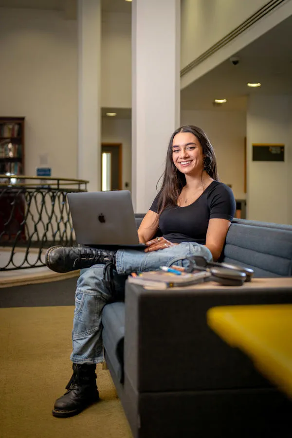 Philosophy student Mayla Singh sits on sofa with an open laptop balanced on her crossed leg
