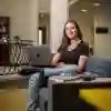 Philosophy student Mayla Singh sits on sofa with an open laptop balanced on her crossed leg