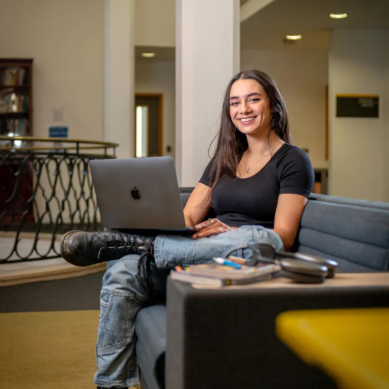 Philosophy student Mayla Singh sits on sofa with an open laptop balanced on her crossed leg