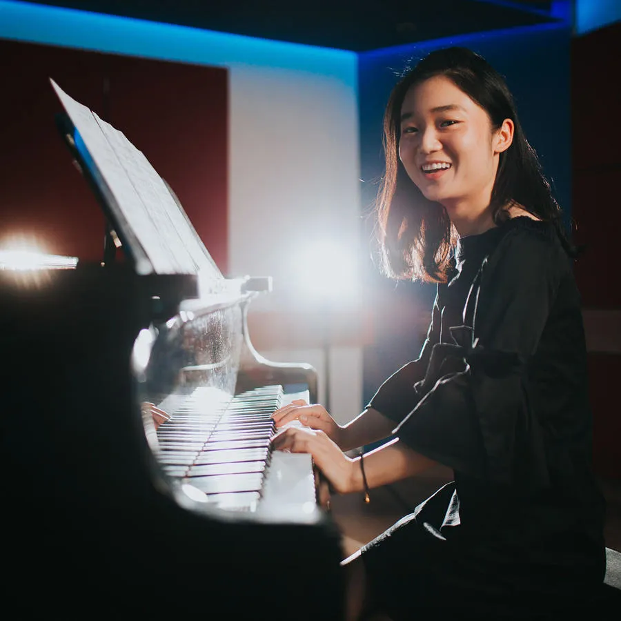 A young woman with her hands on the piano smiles at the camera in a dimly lit room