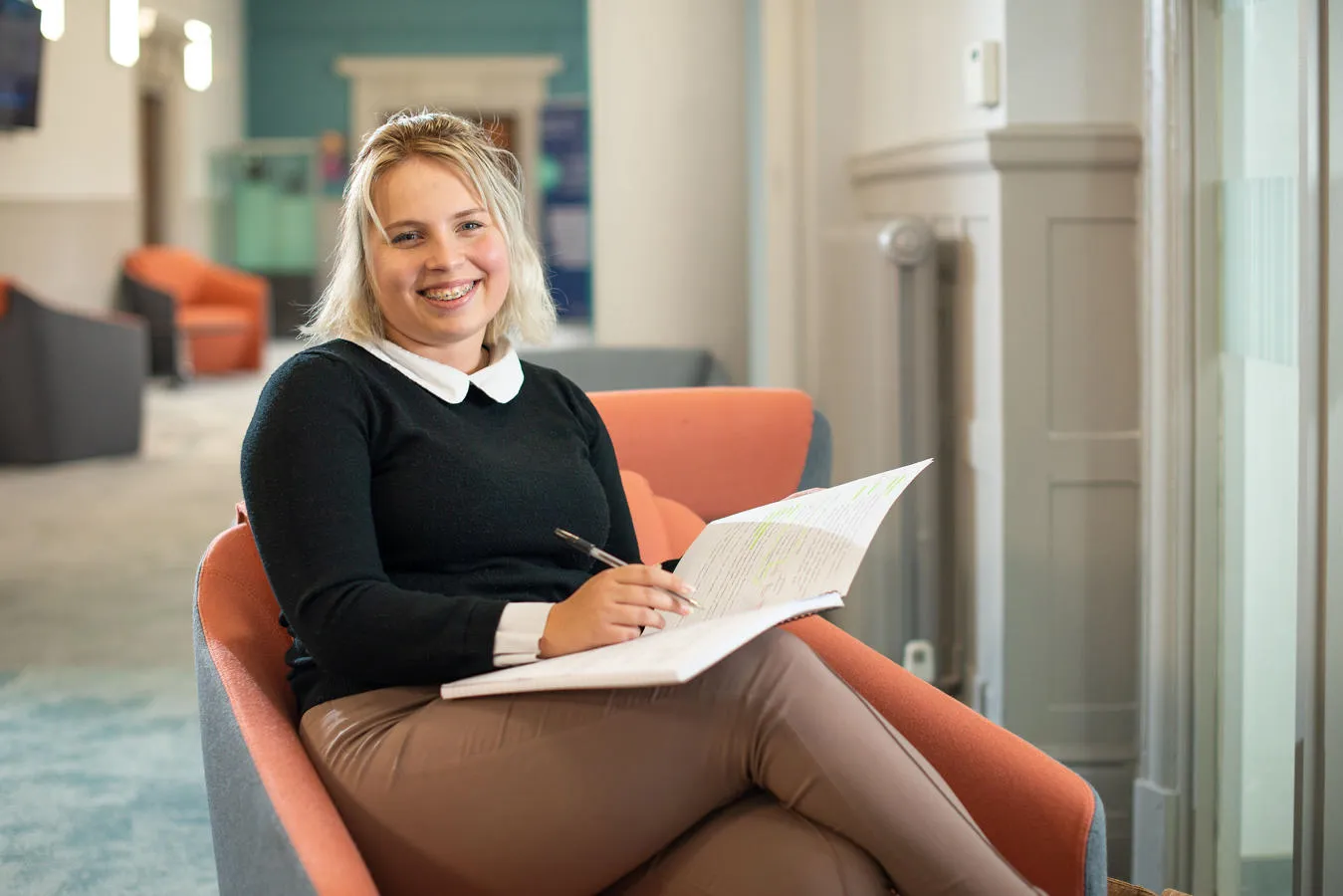 A student seated in an orange chair holds an open notebook and pen