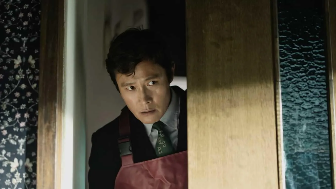 A Korean man in a red apron looks around an open door with a worried look on his face