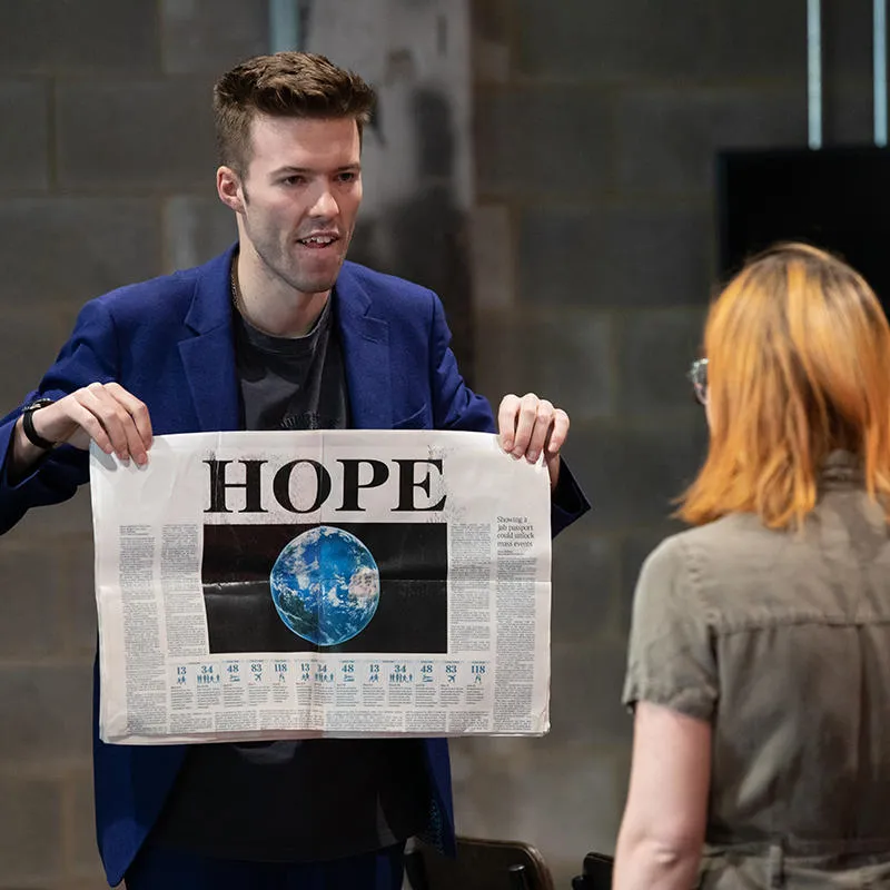 A performer shows a newspaper with headline 'Hope' and image of the Earth