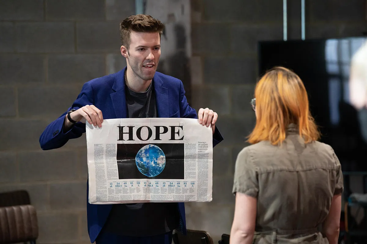 A performer shows a newspaper with headline 'Hope' and image of the Earth 
