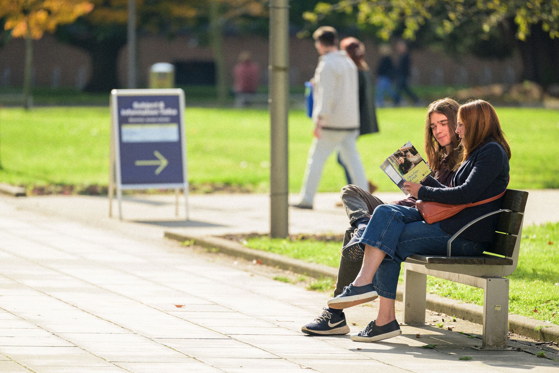 A student and his mum sit on a bench on campus on a sunny day looking at an Open Day guide