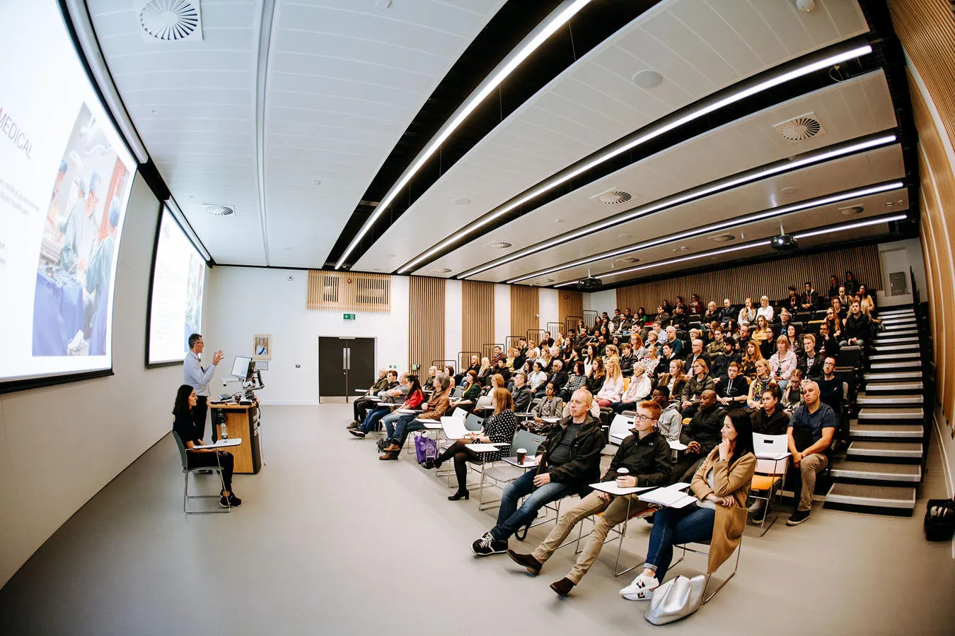 People watch a man give a presentation in a large lecture theatre with screens to the front