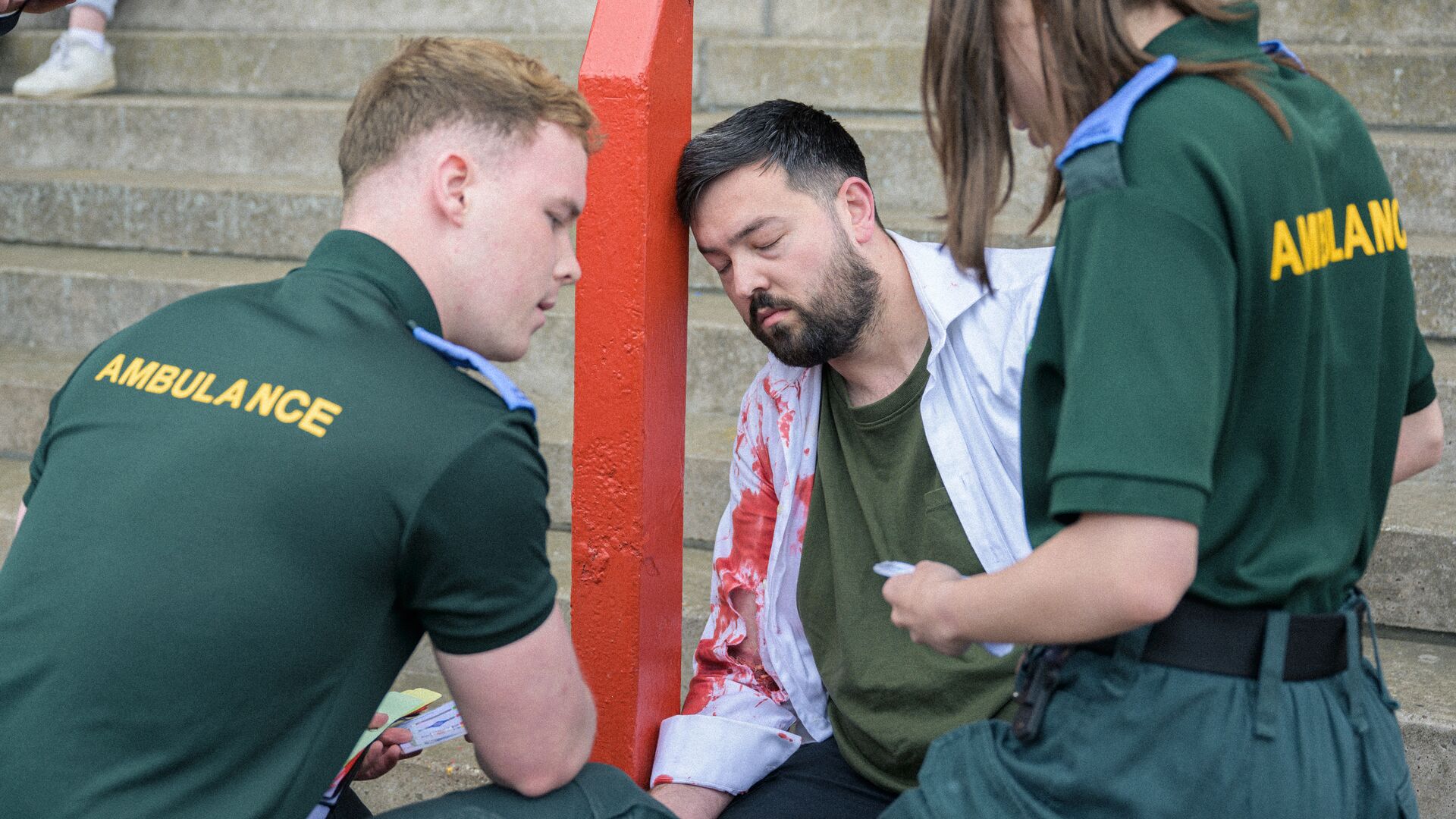Two student paramedics practising on a patient leaning against a red post with blood on his arm