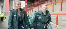 Two student paramedics in green uniforms walking through a sports stadium carrying equipment