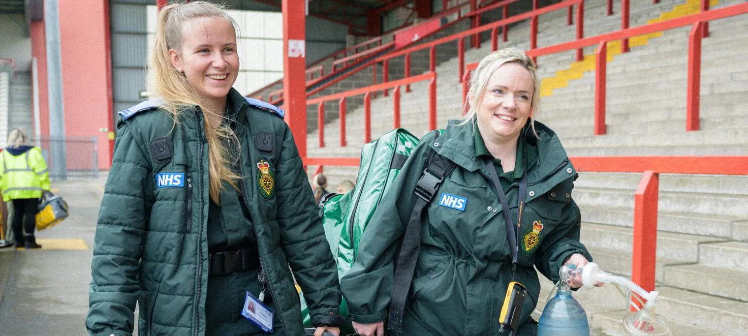 Two student paramedics in green uniforms walking through a sports stadium carrying equipment