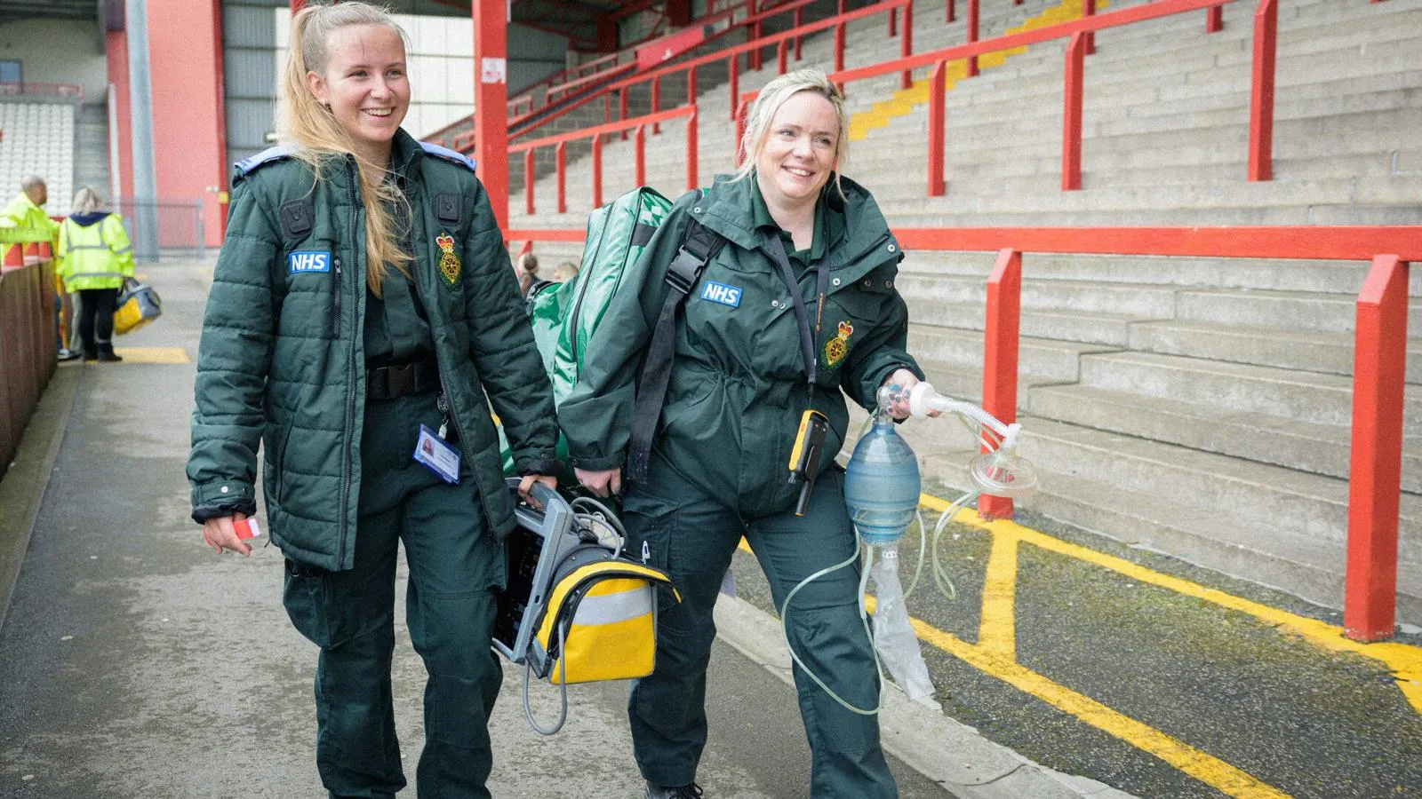 Two student paramedics in green uniforms walking through a sports stadium carrying equipment