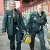 Two student paramedics in green uniforms walking through a sports stadium carrying equipment