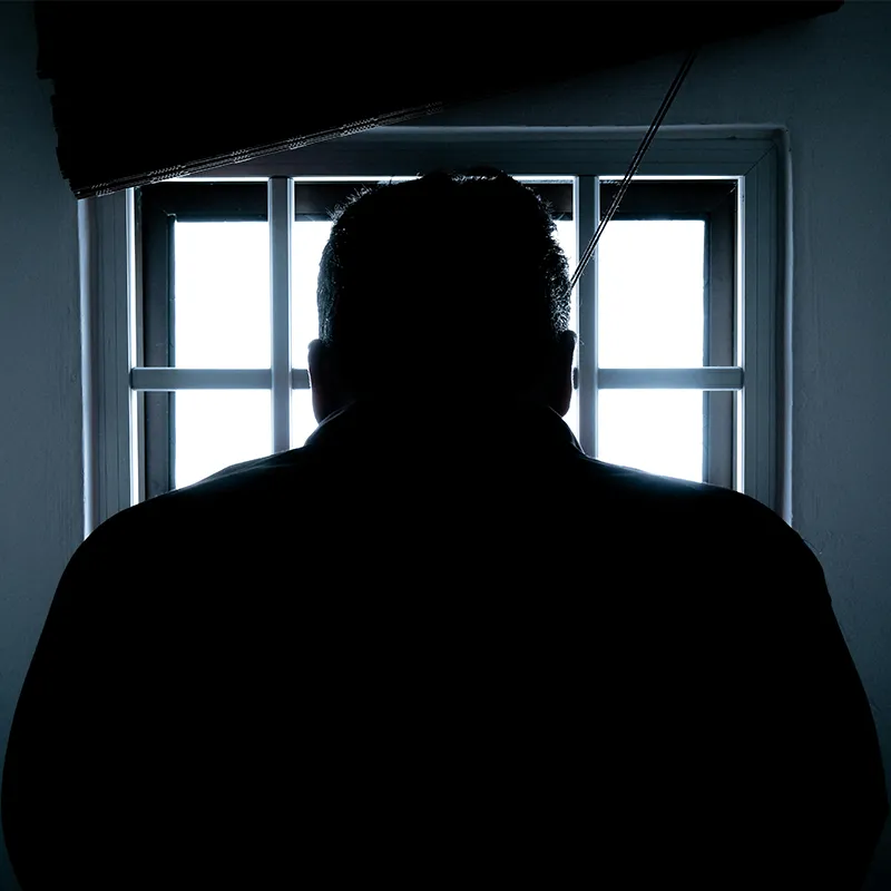 Man in silhouette seen from behind looking through a window with bars 