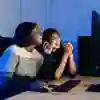 Two young woman chat and look at a computer screen in a dimly lit room