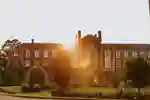 A large historic red-brick building at dusk with a grass lawn and trees and bushes in the foreground.