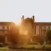 A red-brick building with the sun setting behind. In the foreground is a lawn with trees and bushes surrounding it